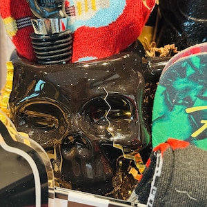 May include: A black skull-shaped container with a decorative bottle stopper. The bottle stopper is black and silver. A red sock with a blue and yellow guitar design is visible. The container is in a gift box with a black and white checkered pattern.