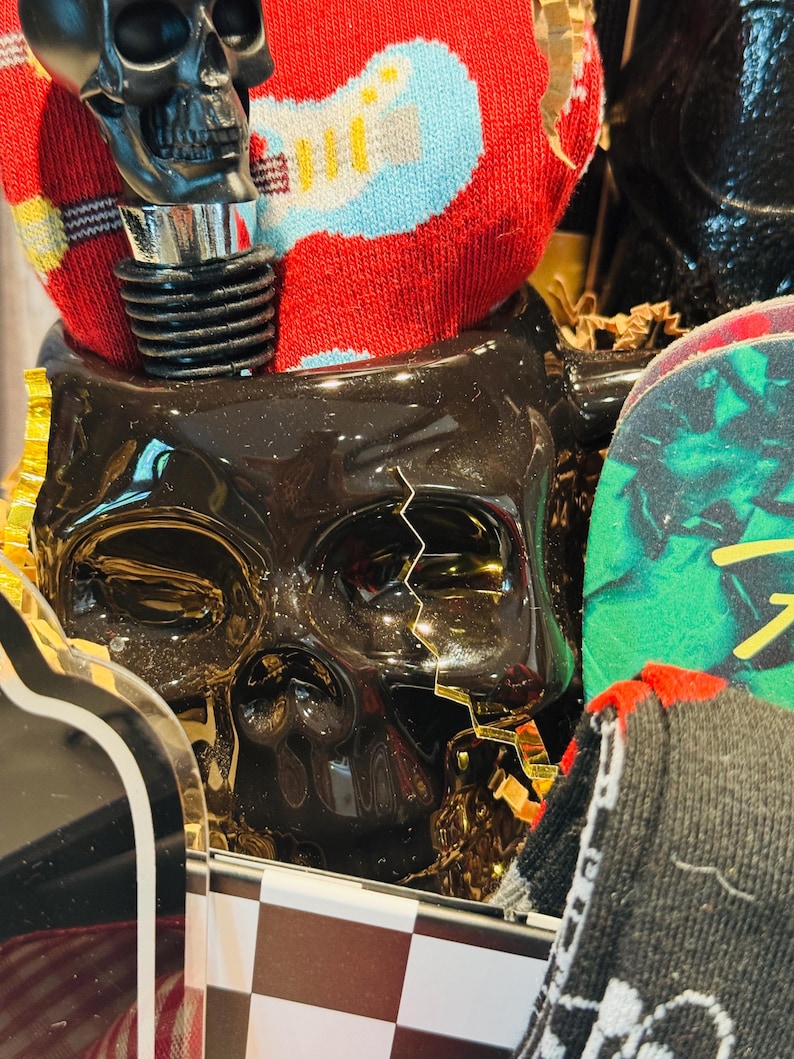 May include: A black skull-shaped container with a decorative bottle stopper. The bottle stopper is black and silver. A red sock with a blue and yellow guitar design is visible. The container is in a gift box with a black and white checkered pattern.
