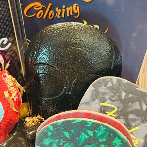 May include: A black, textured skull-shaped object is the focal point, with the word "Coloring" visible in orange. Guitar picks with the Fender logo are in the foreground. The image suggests a theme of music and art.
