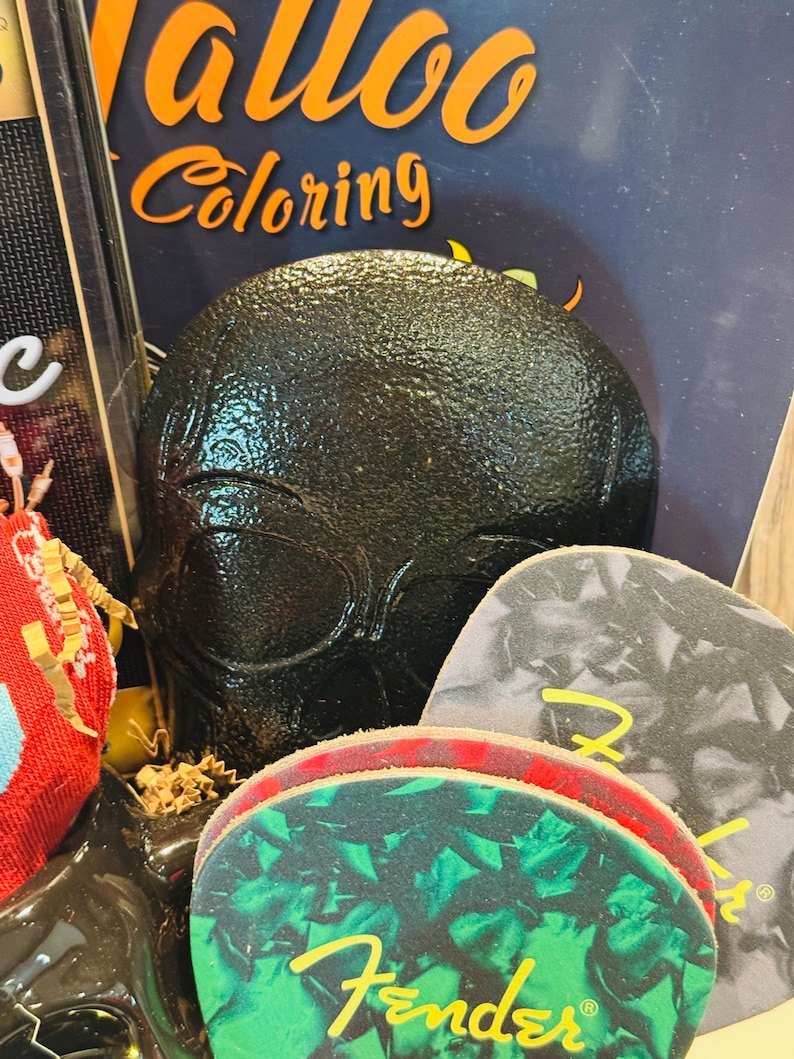 May include: A black, textured skull-shaped object is the focal point, with the word "Coloring" visible in orange. Guitar picks with the Fender logo are in the foreground. The image suggests a theme of music and art.