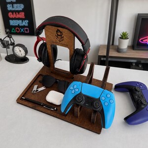 May include: A dark brown wooden gaming organizer stand featuring a vertical headphone mount with a logo, a tray for accessories, and a dedicated slot for a blue and black game controller. A second purple and black game controller sits nearby.
