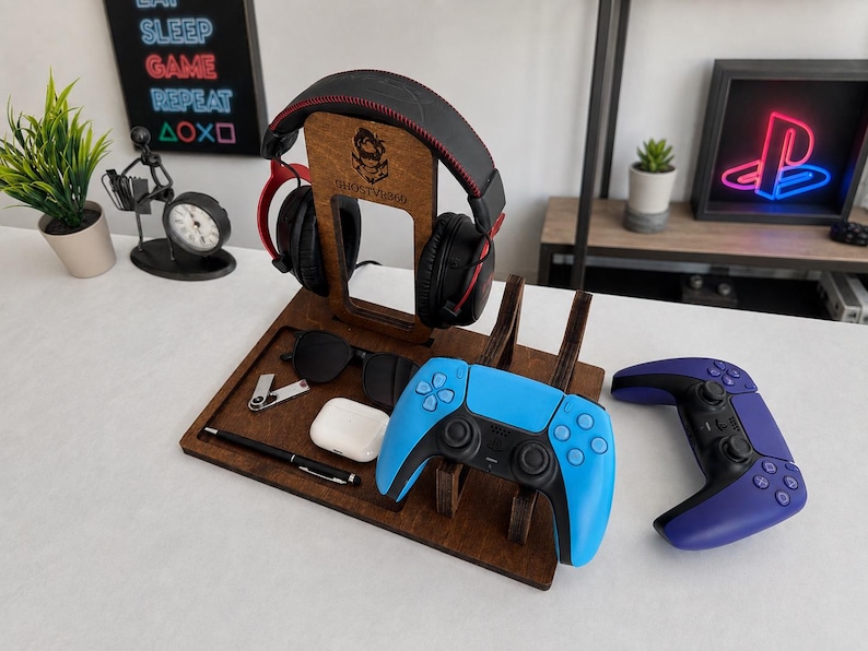 May include: A dark brown wooden gaming organizer stand featuring a vertical headphone mount with a logo, a tray for accessories, and a dedicated slot for a blue and black game controller. A second purple and black game controller sits nearby.