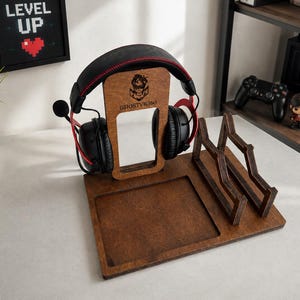 May include: A dark brown wooden gaming organizer stand featuring a vertical headphone hanger with the engraved text GHOSTVR360, a square recessed tray, and a dual-slot controller or accessory holder, currently holding black gaming headphones.