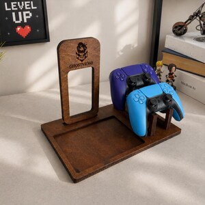 May include: A dark brown wooden gaming controller stand featuring a rectangular base with a recessed tray and an upright support panel. The panel displays a laser-engraved logo and the text GHOSTVR360. Two wireless controllers are mounted on the stand.