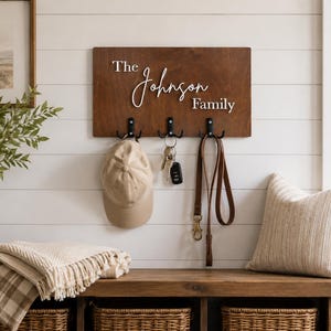May include: A rectangular dark brown wooden wall-mounted hook rack featuring the white 3D text The Johnson Family. The board includes three black metal double hooks holding a beige baseball cap, a set of silver keys, and a brown leather leash.