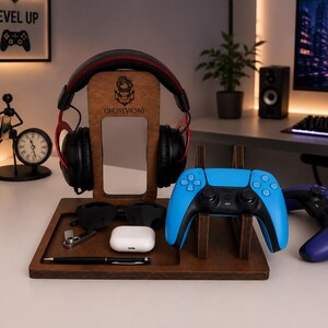 May include: A dark brown wooden gaming organizer stand featuring a central headphone hanger with the engraved text GHOSTVR360, a dedicated slot for a blue game controller, and a flat tray base holding black sunglasses, a white earbud case, and a pen.