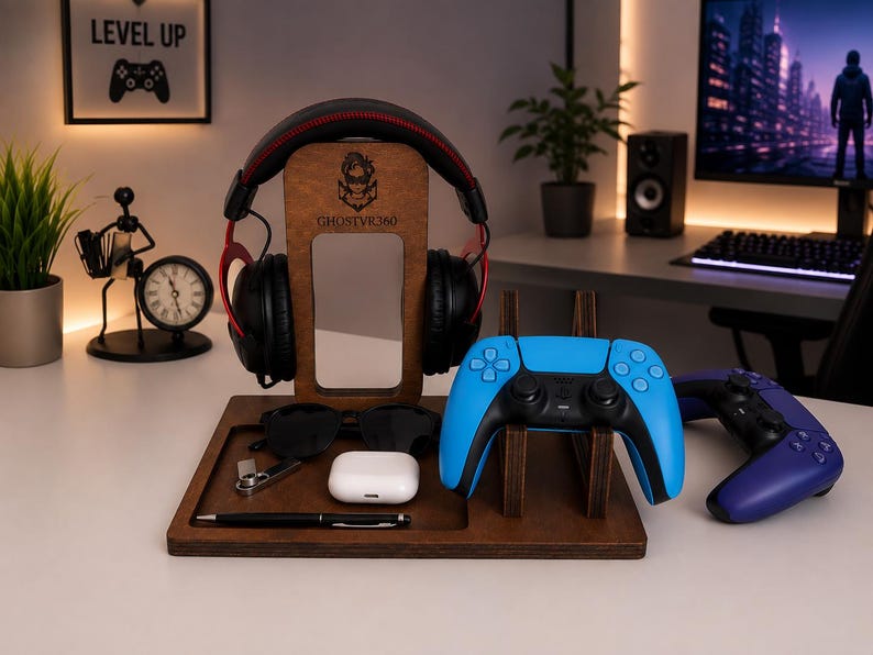 May include: A dark brown wooden gaming organizer stand featuring a central headphone hanger with the engraved text GHOSTVR360, a dedicated slot for a blue game controller, and a flat tray base holding black sunglasses, a white earbud case, and a pen.