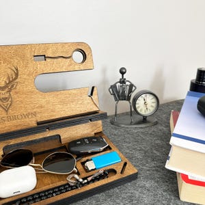 May include: A wooden phone stand with a deer design and the name "S. BROWN" engraved. It has slots for a phone, watch, keys, and other accessories. The stand is on a table with a clock, a metal figurine, and books.