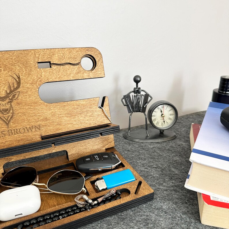 May include: A wooden phone stand with a deer design and the name "S. BROWN" engraved. It has slots for a phone, watch, keys, and other accessories. The stand is on a table with a clock, a metal figurine, and books.