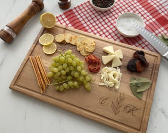 May include: A wooden cutting board with a personalized inscription "E & K" with a leafy design. The board is filled with various snacks, including crackers, grapes, cheese, dried fruit, and breadsticks.