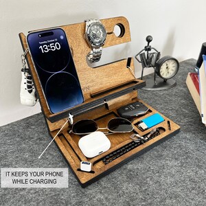 May include: A wooden organizer with compartments for a phone, watch, keys, sunglasses, and other small items. The organizer is brown with black accents and has a slot for charging a phone. The text "Tuesday, October 22" and "13:50" is displayed on the phone screen.