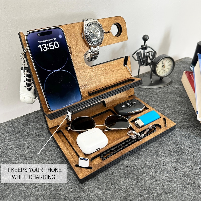 May include: A wooden organizer with compartments for a phone, watch, keys, sunglasses, and other small items. The organizer is brown with black accents and has a slot for charging a phone. The text "Tuesday, October 22" and "13:50" is displayed on the phone screen.