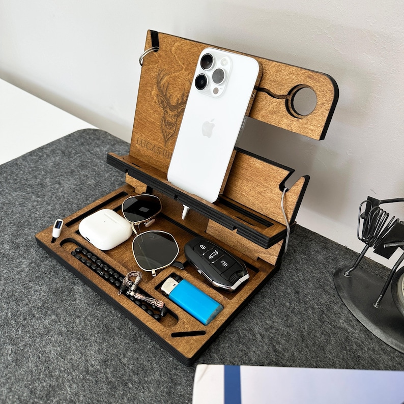 May include: A wooden phone stand with a personalized inscription "Lucas Hill" and a deer head design. The stand has slots for a phone, keys, sunglasses, and a lighter.