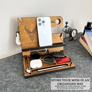 May include: Wooden phone stand with a deer design, personalized with the name "LUCAS BROM". The stand has slots for a phone, keys, sunglasses, and a watch. It also has a space for a charging cable.