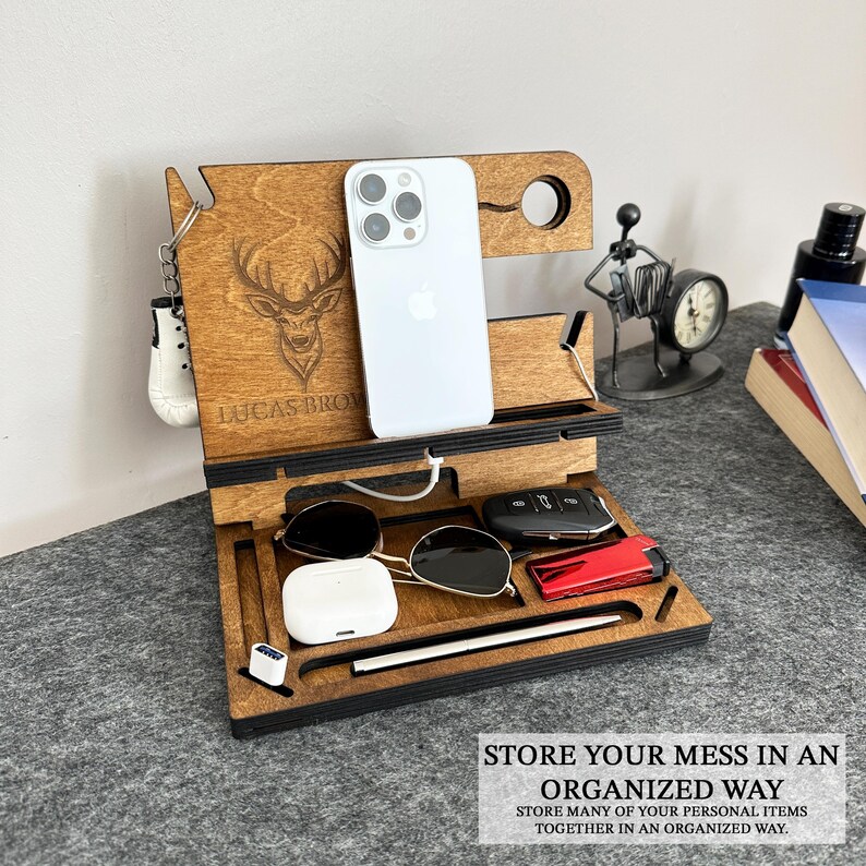 May include: Wooden phone stand with a deer design, personalized with the name "LUCAS BROM". The stand has slots for a phone, keys, sunglasses, and a watch. It also has a space for a charging cable.