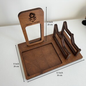 May include: A brown wooden VR headset and controller stand featuring a vertical support with the engraved text GHOSTVR360 and a logo. The base measures 8 inches by 12 inches, with a total height of 8 inches, including a square tray and two cradles.