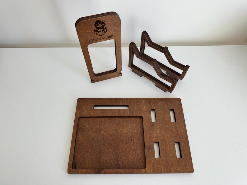 May include: A dark brown wooden organizer set consisting of a rectangular base tray with cutouts, a vertical stand featuring the engraved text GHOSTVR360 and a character icon, and a separate two-tiered rack with angled supports for holding items.