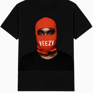 May include: Black t-shirt featuring a person wearing an orange balaclava with the white word "YEEZY" across the front. The person's eyes are visible. The shirt is a crew neck style.