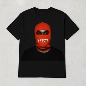 May include: Black t-shirt featuring a person wearing an orange balaclava with the word "YEEZY" in white block letters. The person's eyes are visible through the balaclava's eye opening. The shirt is a classic crew neck style.