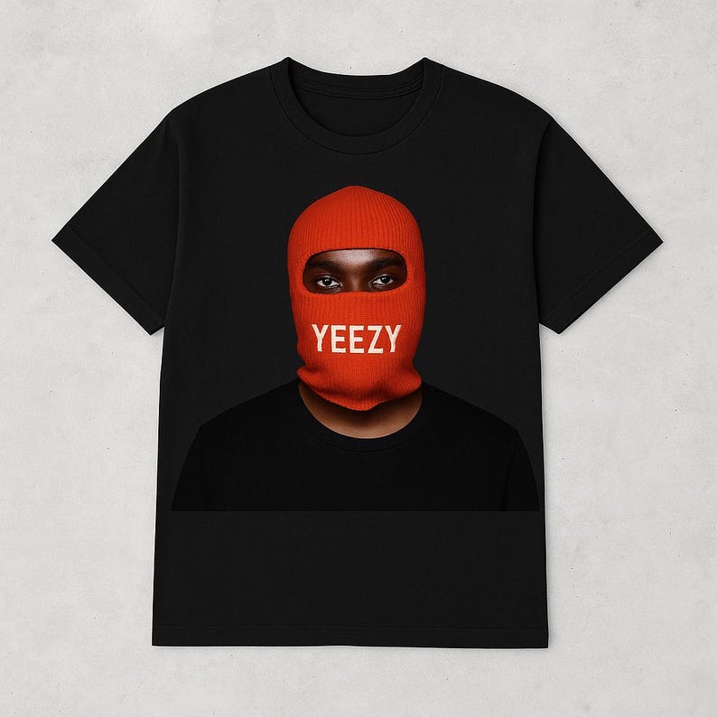 May include: Black t-shirt featuring a person wearing an orange balaclava with the word "YEEZY" in white block letters. The person's eyes are visible through the balaclava's eye opening. The shirt is a classic crew neck style.