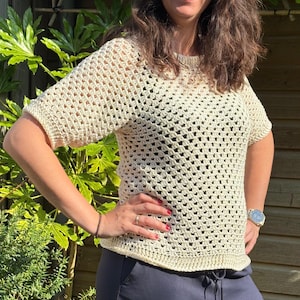 May include: A cream-colored short-sleeved top featuring an open-work crochet pattern with a round neckline and a ribbed hem. The garment is constructed with a repeating square stitch design, providing a textured, semi-sheer appearance throughout.