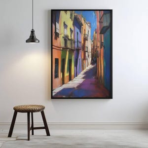 May include: A framed art print depicting a colorful street scene with buildings in shades of orange, yellow, purple, and blue. The artwork captures a sunlit alleyway with shadows and a clear blue sky. The print is displayed on a white wall.