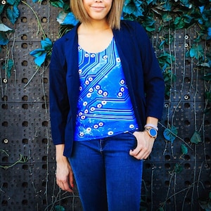 以下が含まれることがあります： 青いブレザー、青と白の模様のシャツ、青いジーンズ、金色の腕時計をつけた女性。彼女は青と緑のツタで覆われた壁の前に立っている。