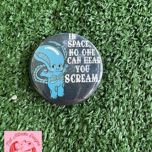 May include: A circular pinback button featuring a dark blue background with white text that reads, IN SPACE, NO ONE CAN HEAR YOU SCREAM. A light blue cartoon creature with a large head and tail is positioned to the left of the white text.