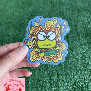 May include: A die-cut vinyl sticker featuring a cartoon green frog character with an angry expression, holding two silver swords. The character is surrounded by stylized orange and yellow flames against a light blue background with a thin white border.