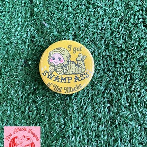 May include: A round yellow pinback button featuring a cartoon creature with green scales and a pink face lying down. The text reads I got SWAMP ASS at Nat Attacks in black lettering. The design includes small white star accents on the yellow background.