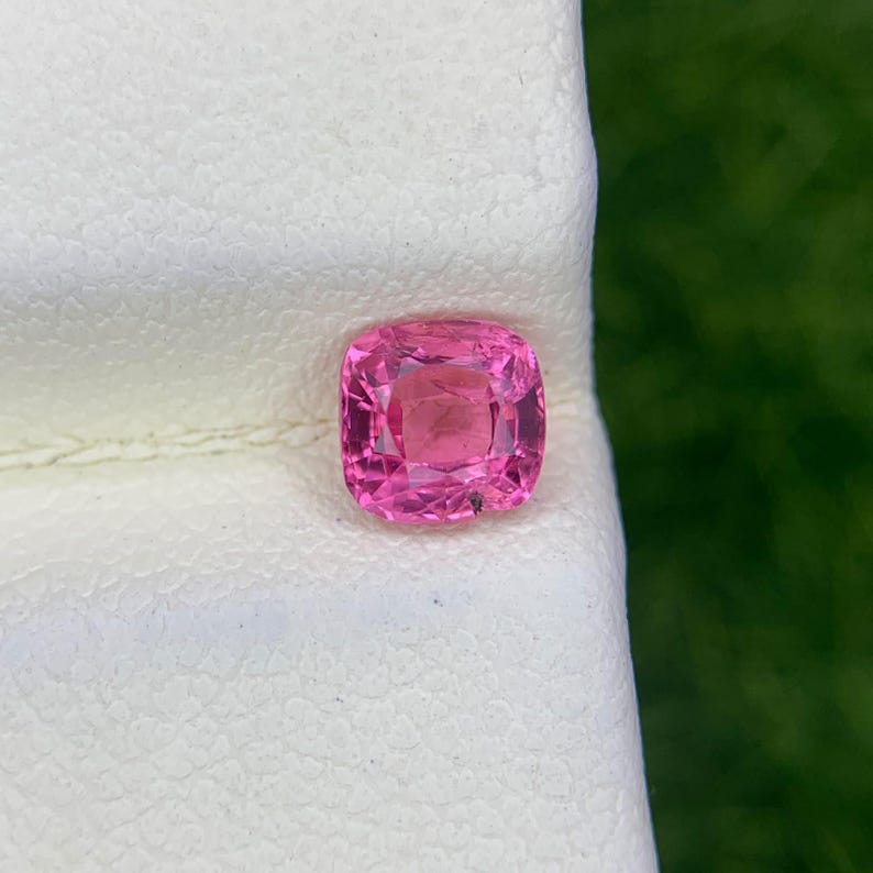 Op de afbeelding: Een kussengeslepen edelsteen met een levendige roze tint en een gefacetteerd oppervlak dat licht reflecteert. De steen ligt op een wit oppervlak met textuur en toont de doorschijnende kwaliteit en de precieze geometrische vorm met facetten.