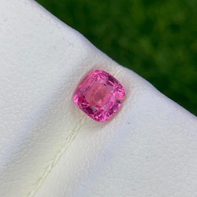 Op de afbeelding: Een levendige, kussengeslepen roze edelsteen met een gefacetteerd oppervlak dat licht reflecteert. De steen vertoont een diepe, verzadigde magentakleur en is geplaatst op een getextureerd, gebroken wit oppervlak met een heldere structuur.