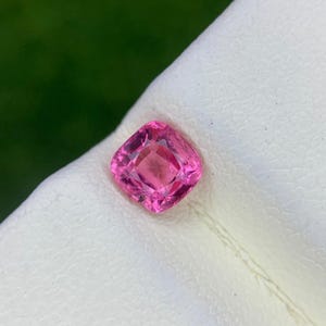 Op de afbeelding: Een roze edelsteen in kussenslijpvorm met een gefacetteerd oppervlak dat licht reflecteert. De steen heeft een levendige, verzadigde roze tint en ligt op een wit oppervlak met textuur. De edelsteen heeft een vierkante vorm met afgeronde hoeken en helderheid.