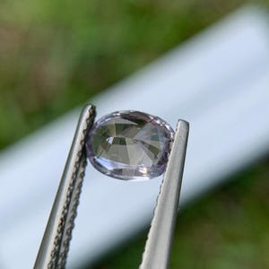 May include: A faceted, oval-shaped gemstone held by tweezers. The gem has a pale lavender hue and is cut to reflect light. The background is blurred, with green and white tones. The tweezers are silver.