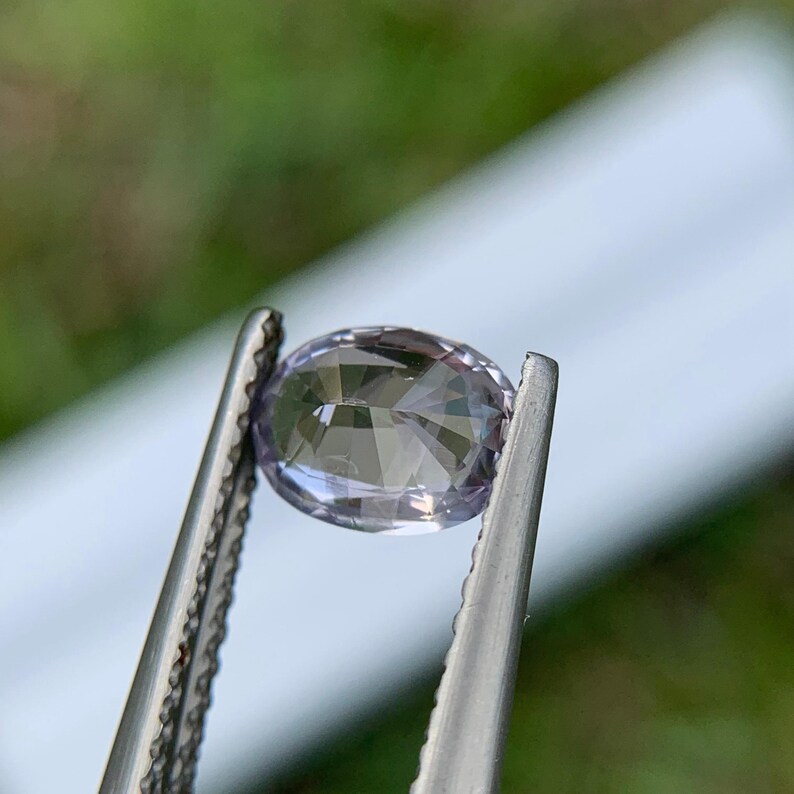 May include: A faceted, oval-shaped gemstone held by tweezers. The gem has a pale lavender hue and is cut to reflect light. The background is blurred, with green and white tones. The tweezers are silver.