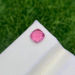 Op de afbeelding: Een roze edelsteen in kussenslijpvorm met een gefacetteerd oppervlak, die een levendige doorschijnende tint vertoont. De steen heeft een symmetrische vierkante vorm met afgeronde hoeken en is geplaatst op een wit, getextureerd presentatieoppervlak.