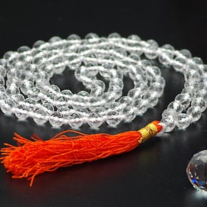 Könnte beinhalten: Eine Mala-Halskette aus klaren Quarz-Kristallperlen mit einer orangefarbenen Quaste und einer facettierten Kristallkugel. Die Perlen sind auf weißem Faden aufgefädelt. Die Mala ist ein spirituelles Werkzeug für Meditation und Gebet.