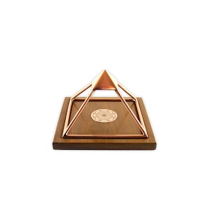 May include: A copper pyramid with a wooden base and a geometric design on the top. The pyramid is on a wooden base with a geometric design.