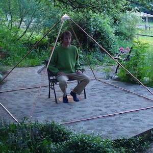 Könnte beinhalten: Eine Person sitzt im Freien in einer Kupferpyramidenstruktur. Die Pyramide besteht aus Kupferrohren und steht auf einer Steinplatte. Die Person trägt einen grünen Pullover und Jeans.