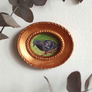 Peut inclure: Un petit cadre ovale doré avec une peinture d'un pigeon gris sur une branche verte à l'intérieur. Le cadre a une bordure perlée.