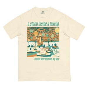 Könnte beinhalten: Cremefarbenes T-Shirt mit einem farbenfrohen Grafikdesign eines Teetassen-Karussells. Der Text "a storm inside a teacup" steht über dem Bild und "shelter here with me, my love" darunter. Das Design verwendet Türkis, Orange und Gelb.