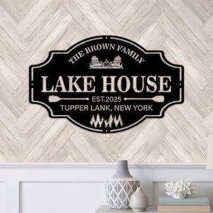 以下が含まれることがあります： 白い文字が入った黒い金属製のレイクハウスの看板。「The Brown Family Lake House」と「Est. 2025」、その下に「Tupper Lank, New York」と書かれています。オール、椅子、木々をあしらった湖をテーマにしています。