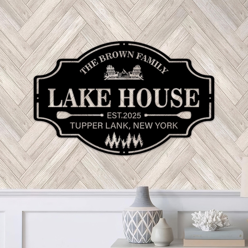 以下が含まれることがあります： 白い文字が入った黒い金属製のレイクハウスの看板。「The Brown Family Lake House」と「Est. 2025」、その下に「Tupper Lank, New York」と書かれています。オール、椅子、木々をあしらった湖をテーマにしています。