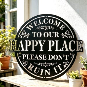 May include: A black metal sign with white lettering that reads "WELCOME TO OUR HAPPY PLACE PLEASE DON'T RUIN IT." The sign is round with decorative flourishes and is displayed outdoors.