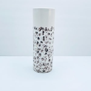 Puede incluir: Un jarrón alto y cilíndrico. La sección superior es blanca, mientras que la sección inferior tiene un patrón texturizado de manchas marrones oscuras y negras. Un jarrón decorativo para el hogar.