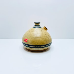 May include: A tan ceramic pitcher with a cork spout and dark blue stripes. The pitcher has a rounded body and a narrow neck with a dark brown top. A small red and white sticker is on the side.