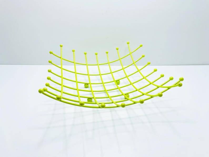 Pu&ograve; includere: Un cestino di frutta in metallo verde lime con un design a griglia. Il cestino ha una forma curva con estremit&agrave; arrotondate e piccoli accenti a sfera. La griglia &egrave; composta da linee sottili e dritte.