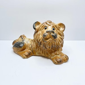 Puede incluir: Figura de león de cerámica en posición reclinada. El león tiene una melena marrón, detalles marrones y negros y una expresión amigable. La figura es un objeto decorativo.