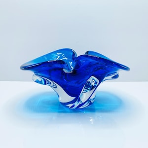 May include: A striking blue glass bowl with an unusual, flowing form. The bowl has a swirling pattern of deep blue and clear glass, producing a captivating visual impact. The edges of the bowl curve outwards, enhancing its artistic quality.