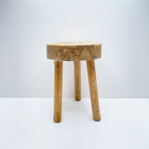 May include: A small, wooden stool with a round top and three legs. The stool is made of light-coloured wood and has a rustic appearance.